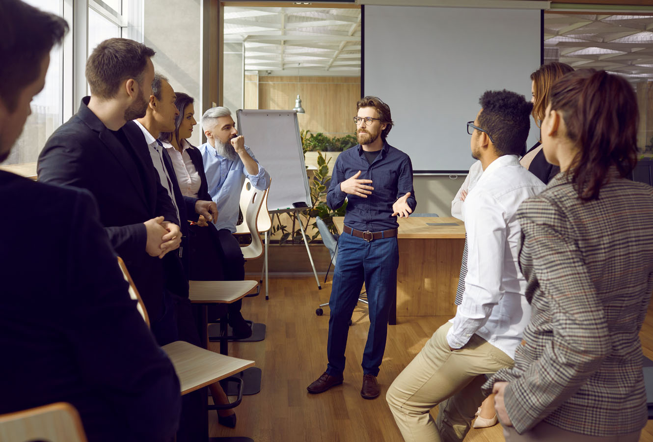 Serious young man business trainer talking to group of people in office standing around him and listening. Office staff, entrepreneurs on corporative training, team building seminar or master class.